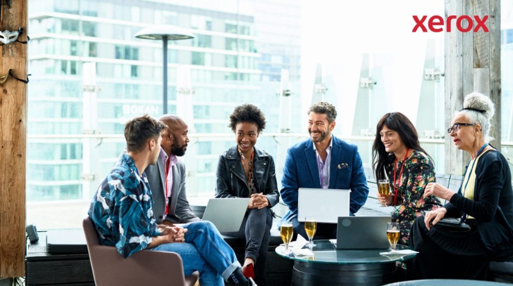 A group of seven people sit together, conversing and working on laptops with a city backdrop A group of seven people sit together, conversing and working on laptops with a city backdrop