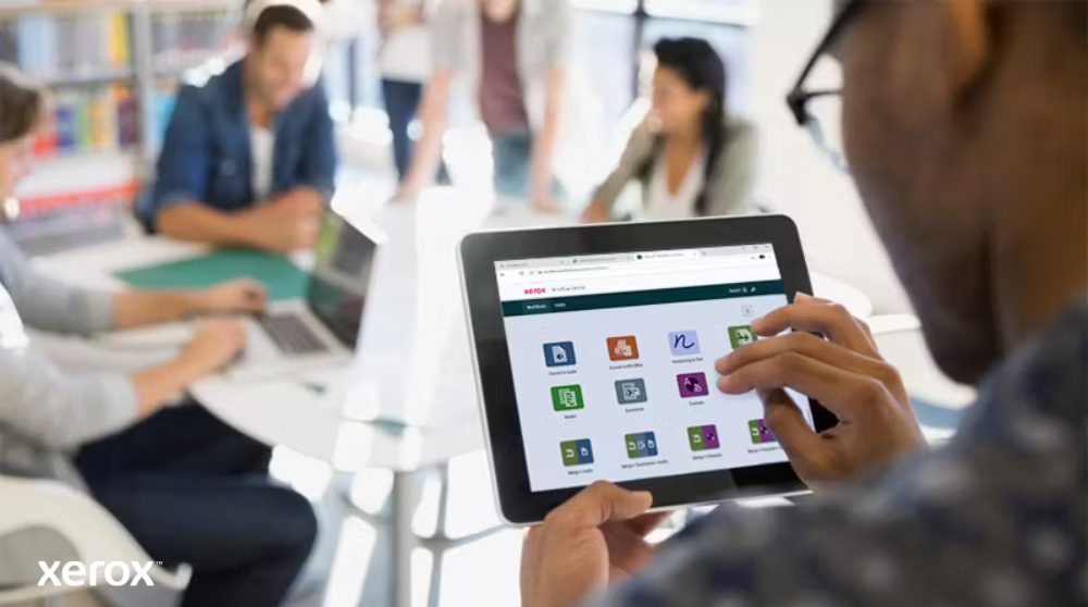 A person holds a tablet displaying the Xerox Workflow Central interface with various app icons, while other individuals work in the blurred background, highlighting mobile access to workflow solutions.
