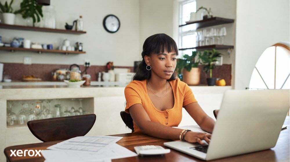 Xerox® DocuShare® Go: Empowering Remote Work A person is sitting at a kitchen table, focused on working on a laptop, with papers and a calculator nearby, illustrating the flexibility of working from home with efficient document management.