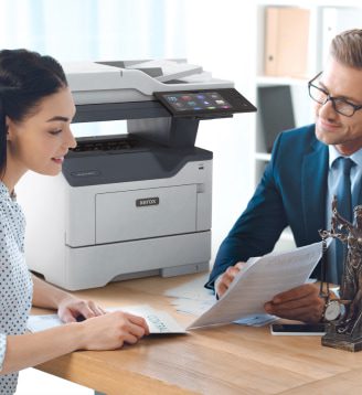 A man and woman seated at a table, engaged in conversation while a Xerox® printer is positioned nearby. A man and woman seated at a table, engaged in conversation while a Xerox® printer is positioned nearby.