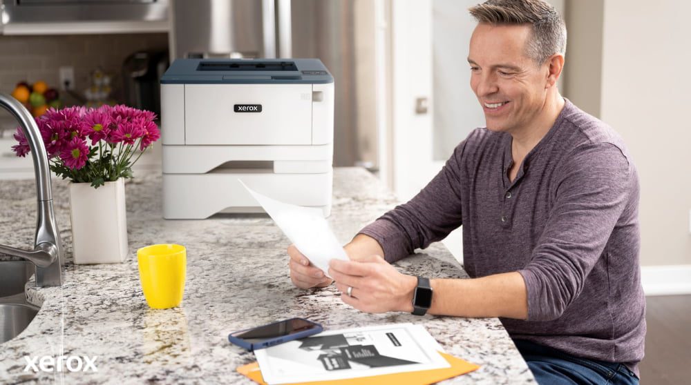 Xerox® Print and Scan Experience: Effortless Document Handling A person is smiling while reviewing a document at a kitchen counter, with a compact Xerox printer and a bright yellow mug nearby, suggesting seamless printing and document handling in a home or small office setting.