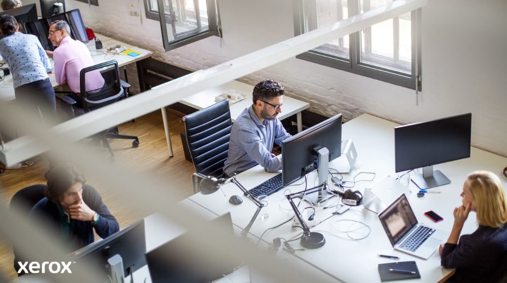 Xerox® Print and Scan Experience: Boosting Office Productivity An overhead view of an open-plan office space with several people working at their desks, using computers. The scene conveys a productive and collaborative work environment.