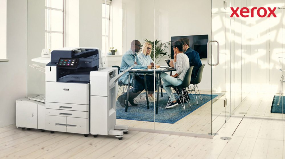 Xerox® Smart Start: Seamless Office Integration A modern Xerox AltaLink B8245 multifunction printer stands in the foreground of a bright office. In the background, a group of people are engaged in a collaborative discussion around a table.