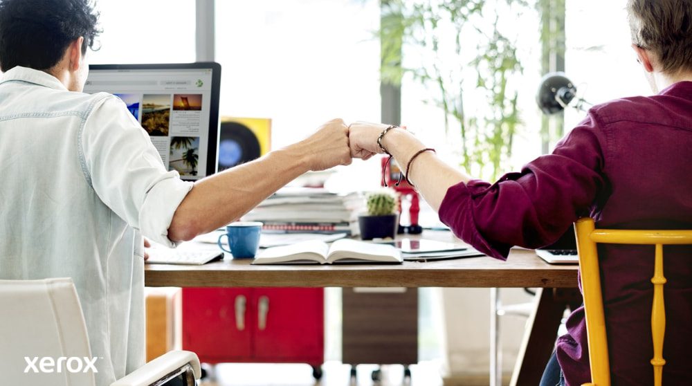 Xerox® Smart Start: Teamwork and Collaboration Two people seated at a desk, facing away from the camera, perform a fist bump, symbolizing teamwork and successful collaboration. One is looking at a computer screen.