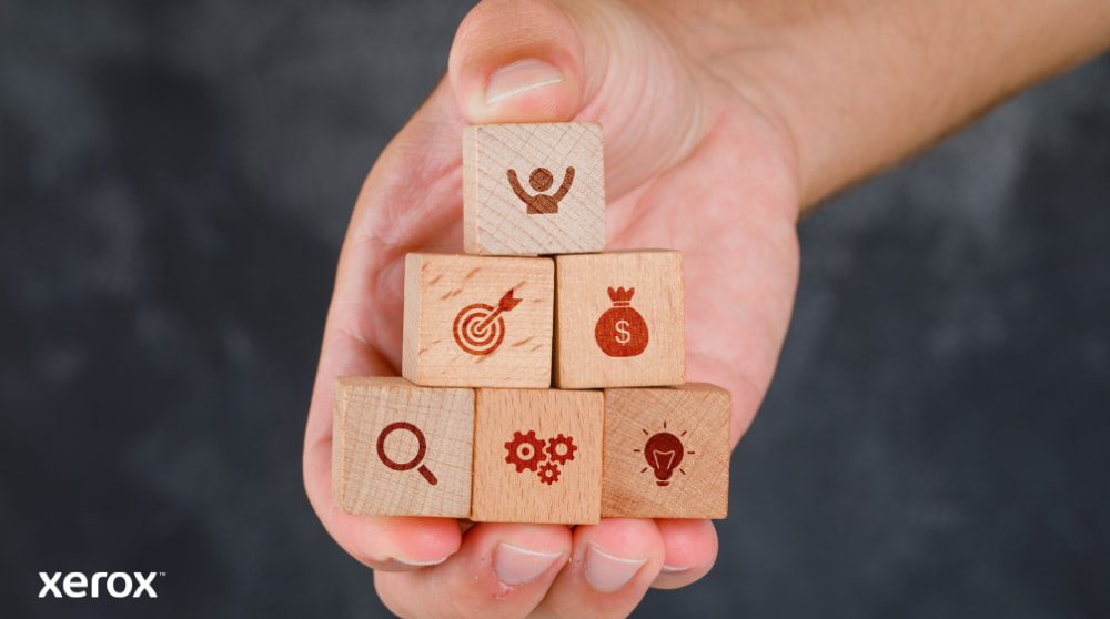 Xerox® Smart Start: Unlocking Business Potential A hand holds a stack of wooden blocks, each engraved with a business icon: a person, a target, a money bag, a magnifying glass, gears, and a lightbulb, representing various benefits and solutions.