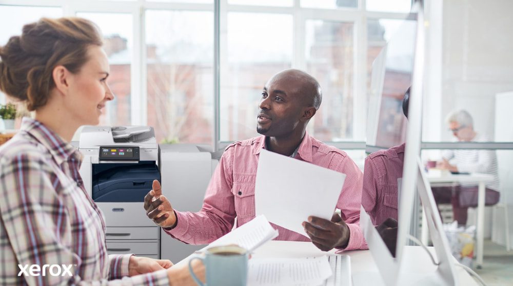 Xerox® Workplace Solutions: Centralized Fleet Management A person reviews data on a laptop screen, with a digital overlay of various device icons (printers, mobile devices) and connected lines, symbolizing centralized fleet management for Xerox Workplace Solutions.