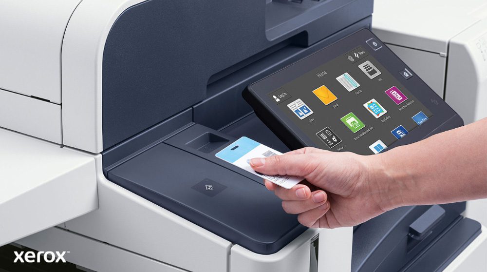 A hand holds a security card, scanning it near the card reader of a modern Xerox multifunction printer with a large touchscreen display showing various application icons.