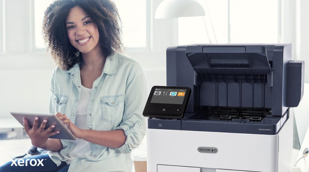 A smiling person is seated next to a white Xerox VersaLink B610 printer, holding a tablet and looking at the camera, highlighting the user-friendly nature of Xerox solutions.
