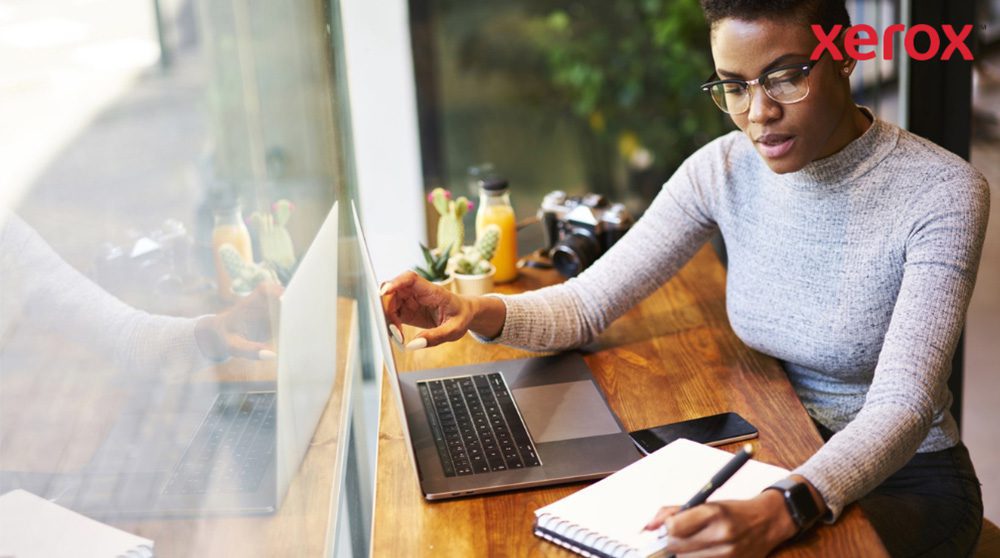A person wearing glasses is working at a wooden table, looking at a laptop screen and writing in a notebook, with a smartphone nearby, suggesting a productive work environment