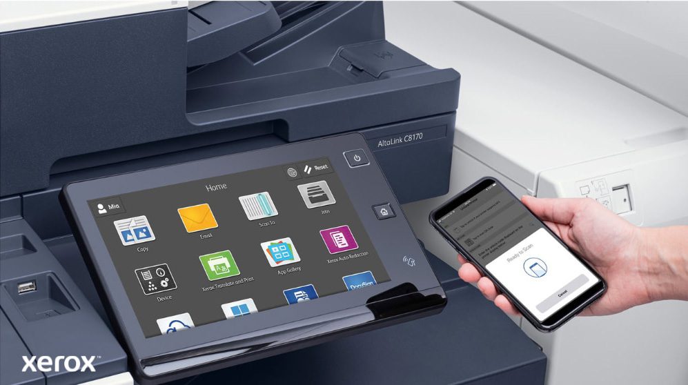 A close-up of a person's hand holding a smartphone, which is wirelessly connected to the touchscreen control panel of a Xerox multifunction printer, showcasing the seamless mobile printing and scanning capabilities of Xerox ConnectKey.