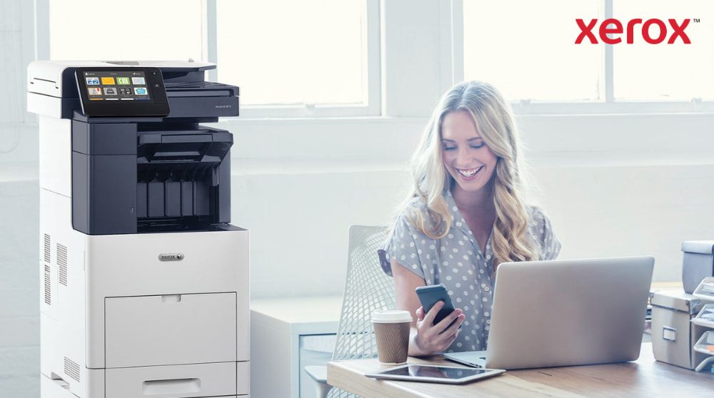 A person is happily looking at their smartphone while sitting at a desk with a laptop. A Xerox VersaLink B605 multifunction printer is visible next to the desk in a brightly lit office space.
