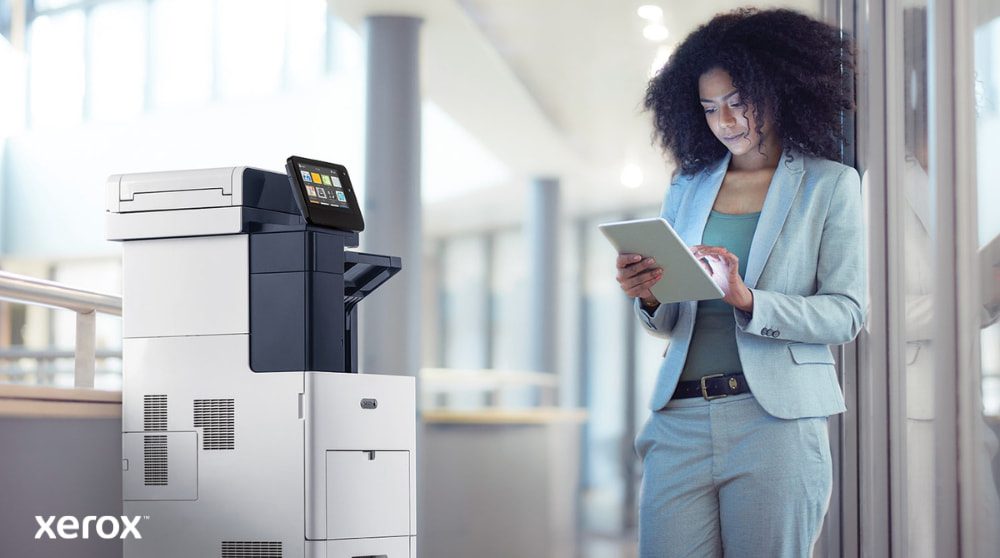 A person stands next to a Xerox VersaLink B605 multifunction printer, looking at and interacting with a tablet device, illustrating how ConnectKey enables mobile interaction with the printer.