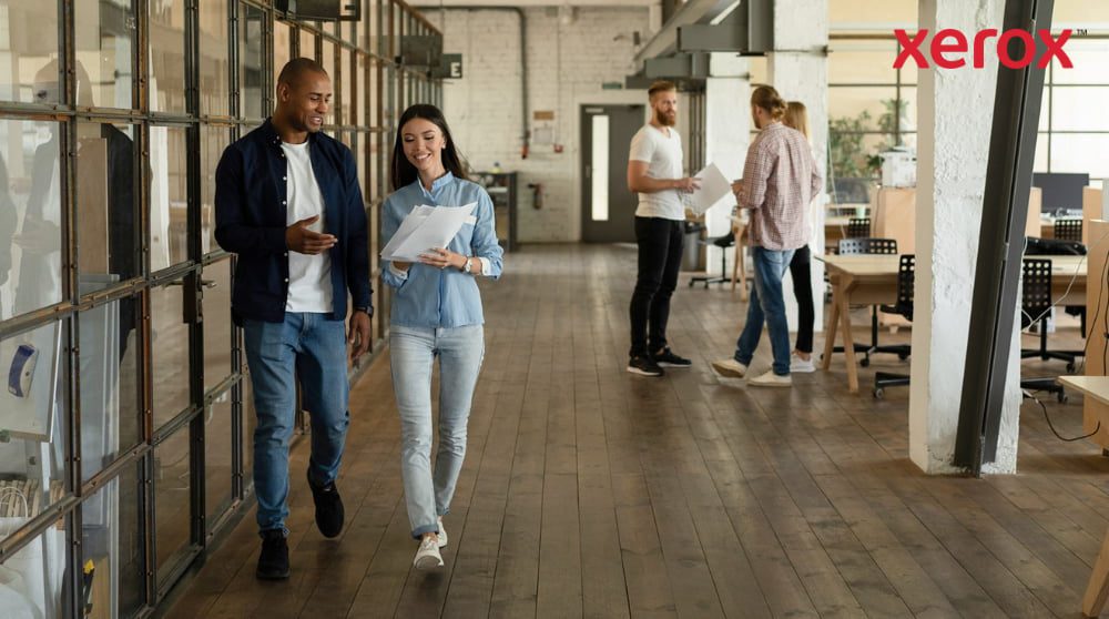 Two colleagues are walking down a hallway in a modern, open-plan office, holding documents and smiling, with other colleagues visible in the background, representing a dynamic and collaborative workplace.