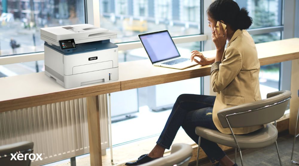 A person is sitting at a high table by a large window, talking on a phone while working on a laptop. A Xerox B235DNI multifunction printer is conveniently placed on the table next to them, suggesting ease of use in a modern workspace.