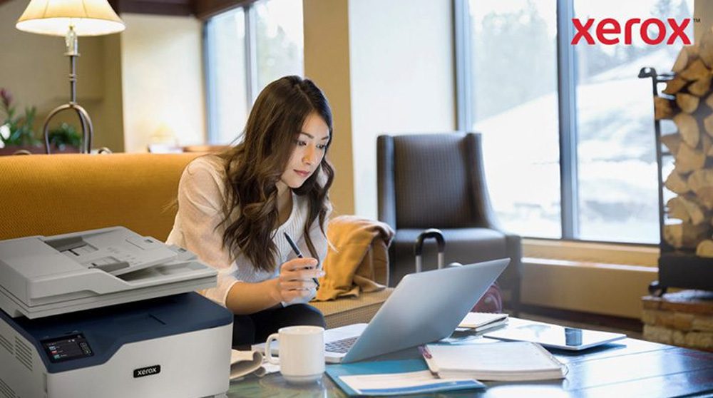 A woman is working on a laptop in a comfortable office or lounge setting, with a compact Xerox printer visible in the foreground, indicating seamless integration of printing with digital workflows.